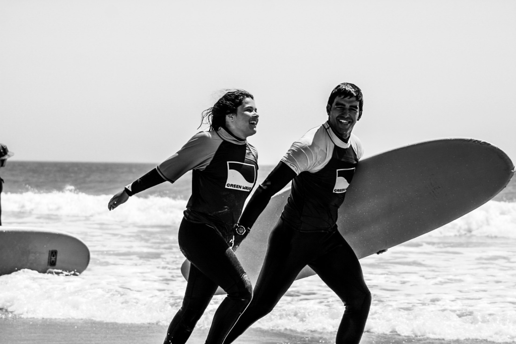 Dois alunos de surf da Green Wave a sorrir, com prancha, a sair da água numa praia em Cascais