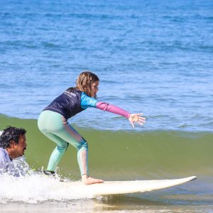 Criança em equilíbrio na prancha de surf sobre uma onda, com instrutor por perto em Cascais